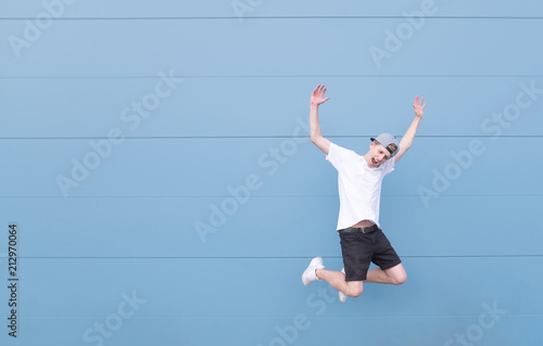 Young man in a white T-shirt and black shorts in a jump on a blue background. Emotional teen jumps against the background of a blue wall