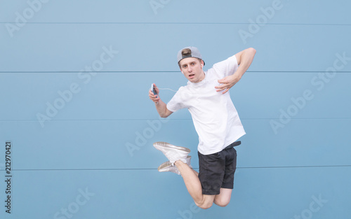 Young man jumps with headphones and music on a pastel blue background. Portrait of a teenager in flight with headphones and a smartphone on a backgraound of a blue wall
