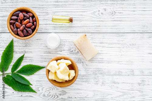 Beautiful composition with cocoa butter cosmetics. Cocoa butter and cocoa beans near homemade soap, cream, oil or lotion in small bottles, green leaves on grey background top view copy space