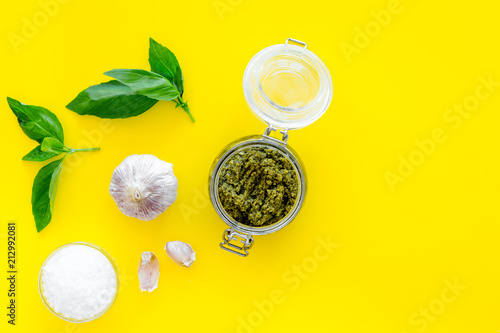 Composition with green pesto sauce in glass jar, basil leaves and garlic on yellow background top view copy space