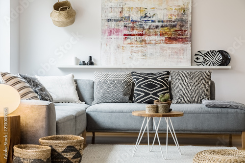 Real photo of a cozy couch with cushions standing behind a small table and in front of a shelf with a painting in boho living room interior with baskets and white rug photo