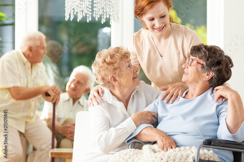 Happy friendship in old age. Tender caregiver standing behind senior women hugging each other in a nursing home. Men in the blurred background.
