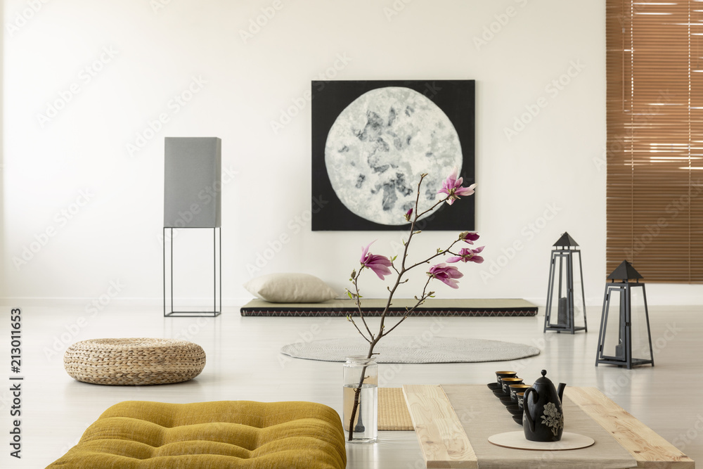 Fototapeta premium Kettle on wooden desk next to yellow pouf and flowers in japanese interior with moon poster. Real photo