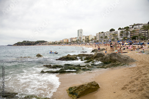 Lloret de Mar / Spain - June 27, 2018: Beach off the coast of Lloret de Mar, Costa Brava. People on the beach lie on sun loungers under umbrellas. Resting under a castle on the beach in the city.