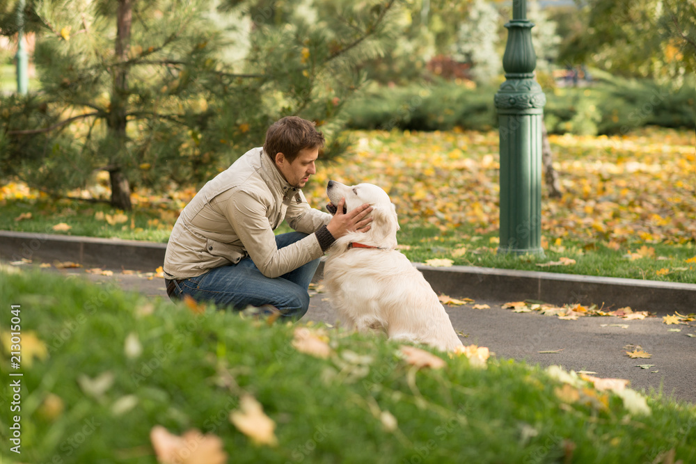 Fototapeta premium The guy wearing biege jacket and jeans is friendly with his dog Labrador in an autumn park.