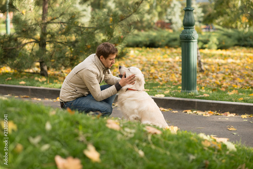The guy wearing biege jacket and jeans is friendly with his dog Labrador in an autumn park. © rozaivn58
