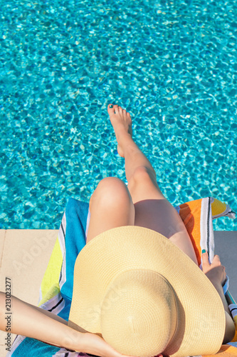 Summertime in pool. Young woman with floppy hat lying in a deck chair by the pool.