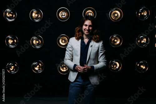 a man in a white jacket and in a black shirt, with long hair, stands against the backdrop of bright lanterns, safites. on the stage.performance. smiles