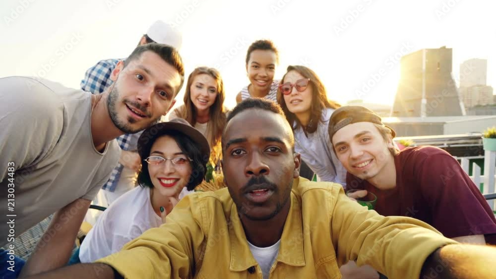 Point of view shot of handsome African American man taking selfie with his happy friends, people are looking at camera, smiling and posing with bottles at roof party. Point of view shot of handsome African American man taking selfie with his happy friends, people are looking at camera, smiling and posing with bottles at roof party.