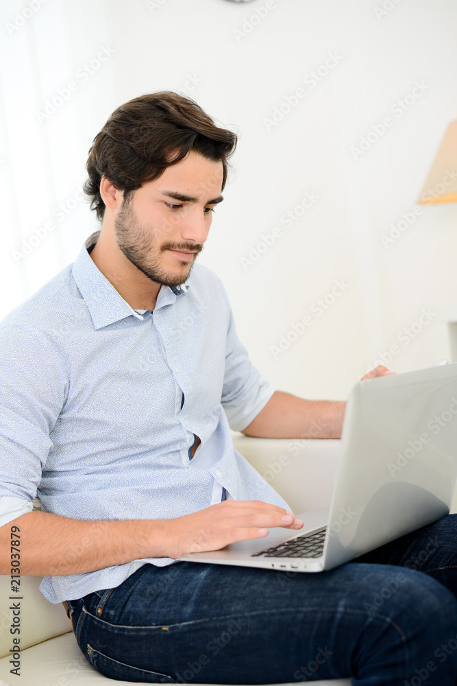 handsome and cheerful young man on sofa at home websurfing and shopping online with laptop computer and internet technology handsome and cheerful young man on sofa at home websurfing and shopping online with laptop computer and internet technology