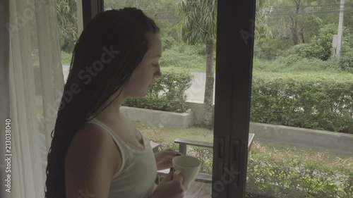 Girl in the morning drinking coffee at a panoramic window overlooking the green garden. S-log, ungraded photo
