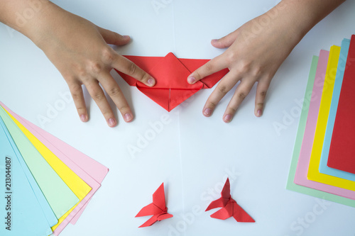 Детские руки делают бабочку оригами из красного листика бумаги на белом фоне. Обучение детей photo