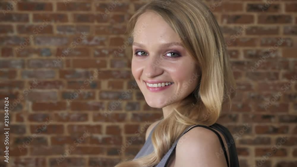 Young cute blond girl is watching at camera, smiling, turn head, brick background, side view Young cute blond girl is watching at camera, smiling, turn head, brick background, side view