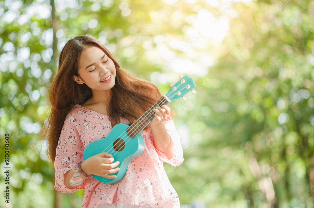 asian lady with hat play ukulele bossanova music in summer time.Young cute  woman playing music outdoors.A happy young girl enjoys playing ukulele  under a tree. Beautiful nature in the background. Stock Photo