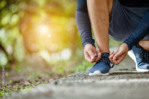 Man tying jogging shoes.A person running outdoors on a sunny day.Focus on a side view of two human hands reaching down to a athletic shoe.Young male jogger athlete training and doing workout outdoors
