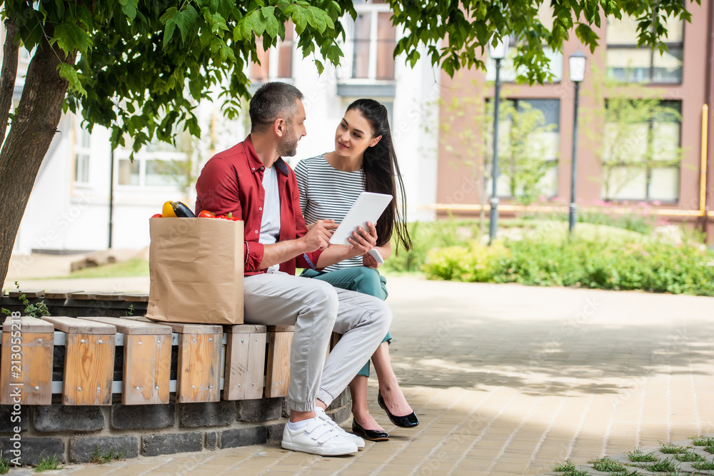 side view of married couple using tablet together while resting after shopping on bench on street side view of married couple using tablet together while resting after shopping on bench on street