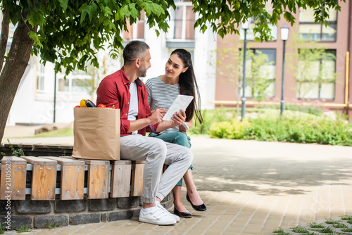 side view of married couple using tablet together while resting after shopping on bench on street