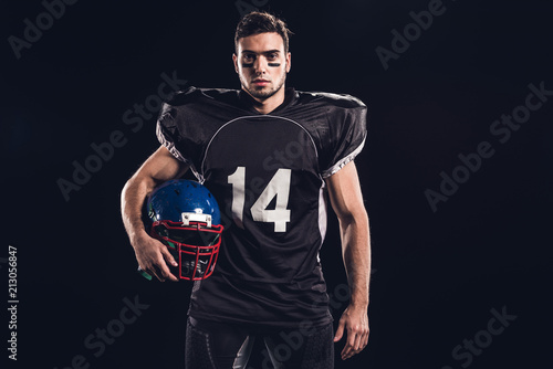young american football player in black uniform holding helmet and looking at camera isolated on black