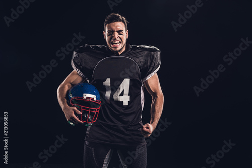 angry american football player in black uniform holding helmet and looking at camera isolated on black