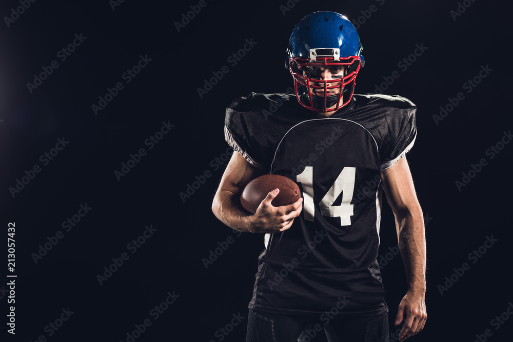 confident american football player holding ball in hand and looking at camera on black
