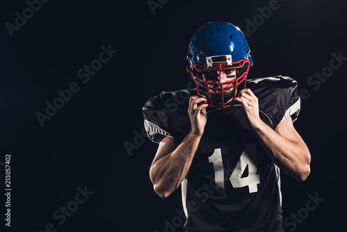 american football player putting on helmet and looking at camera isolated on black