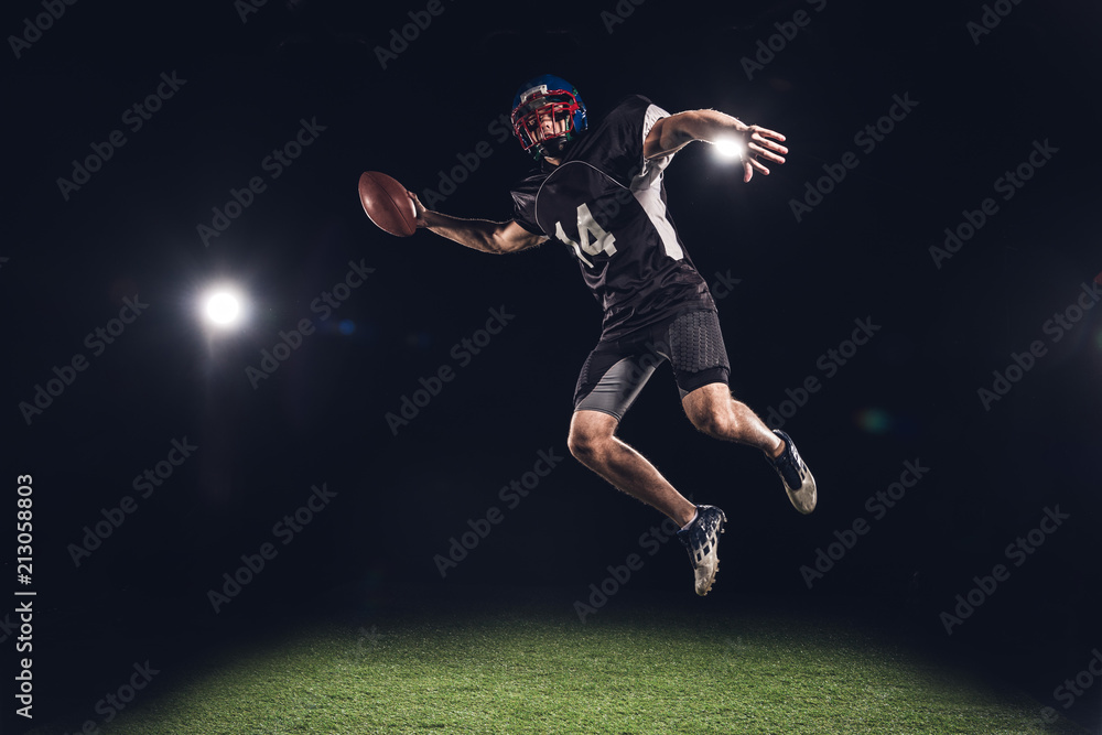 american football player jumping with ball under spotlights on black