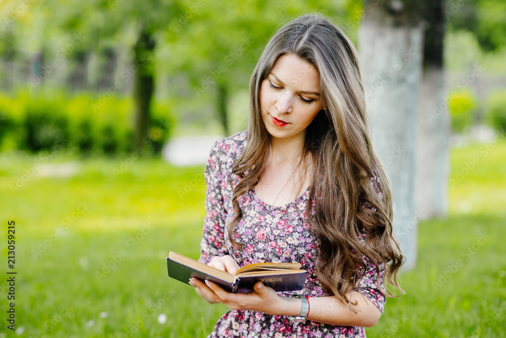 Fototapeta premium Young pretty woman in summer dress relaxing and reading a book in the park