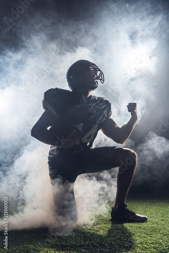 young american football player standing on knee on green grass and raising fist in white smoke