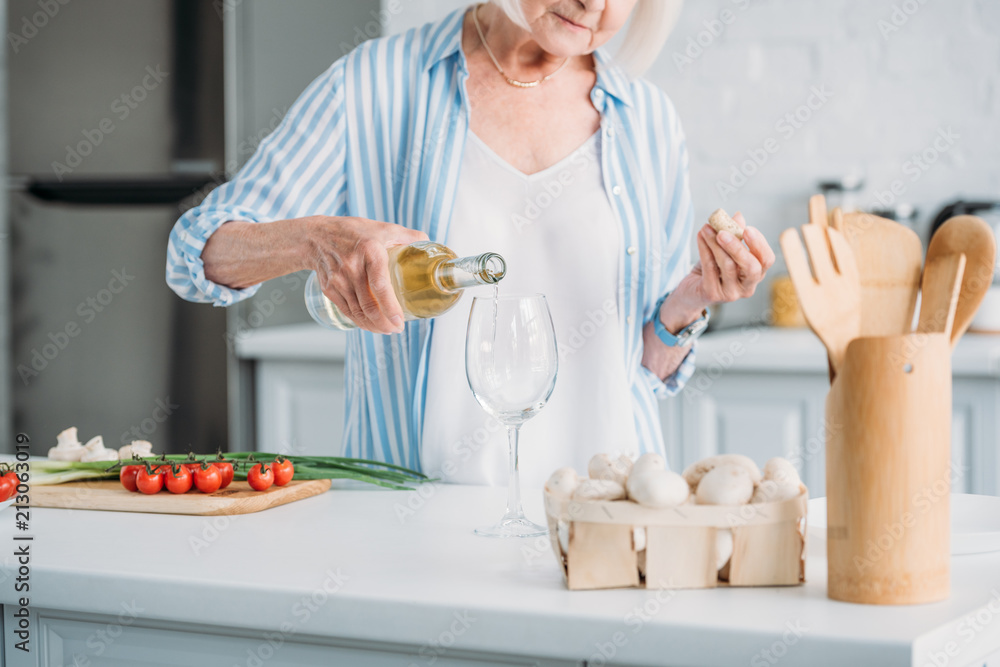 partial view of senior woman pouring wine into glass at counter with fresh vegetables in kitchen partial view of senior woman pouring wine into glass at counter with fresh vegetables in kitchen