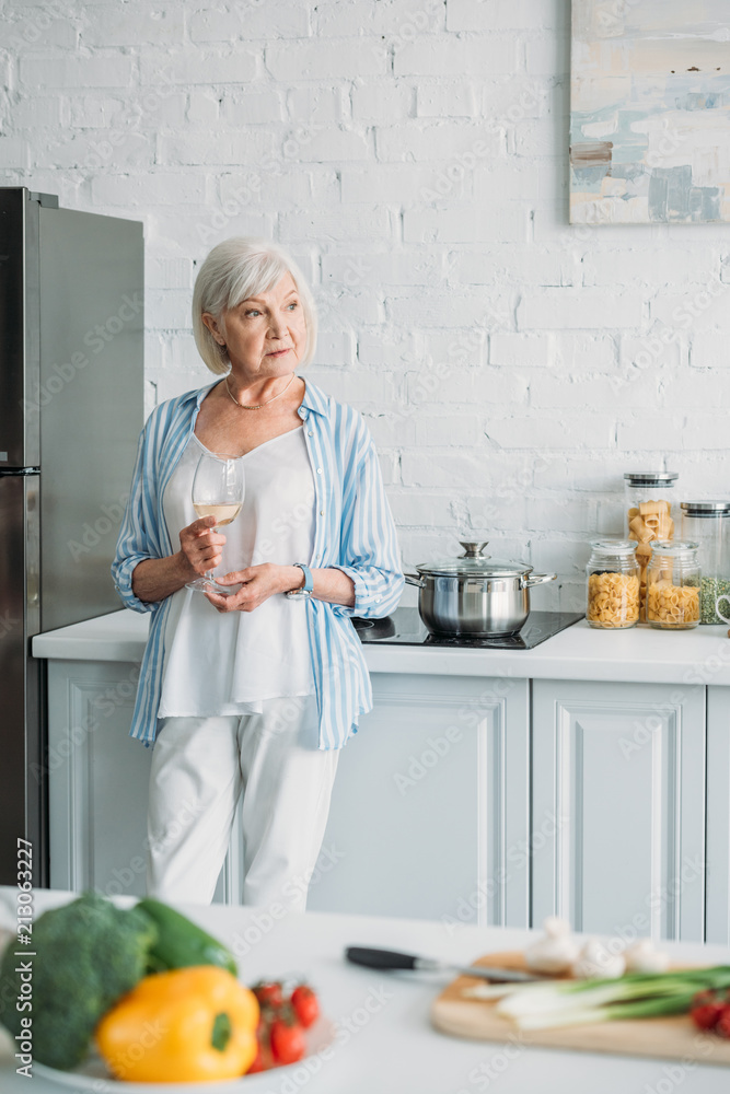 selective focus of fresh vegetables on counter and senior woman with glass of wine standing at stove in kitchen selective focus of fresh vegetables on counter and senior woman with glass of wine standing at stove in kitchen