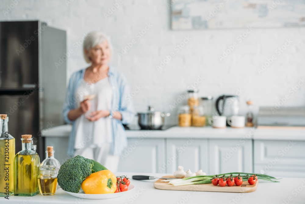 selective focus of fresh vegetables on counter and senior woman with glass of wine standing at stove in kitchen
