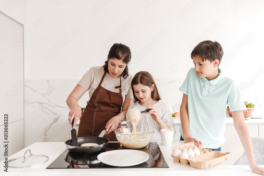 Content woman and happy children 8-10 cooking together, and frying pancakes on modern stove in kitchen at home Content woman and happy children 8-10 cooking together, and frying pancakes on modern stove in kitchen at home