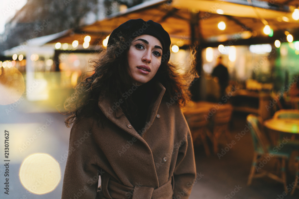 Half length portrait of a beautiful caucasian woman with long dark curly hair wearing stylish coat looking at the camera while standing on a blurred street background. Bokeh light.