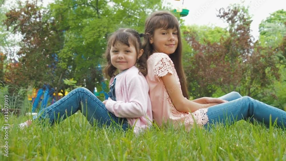 The children are sitting on the grass. Happy little sisters are sitting back to back in the park. The children are sitting on the grass. Happy little sisters are sitting back to back in the park.