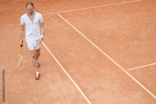 handsome sportsman with tennis racket walking on brown court