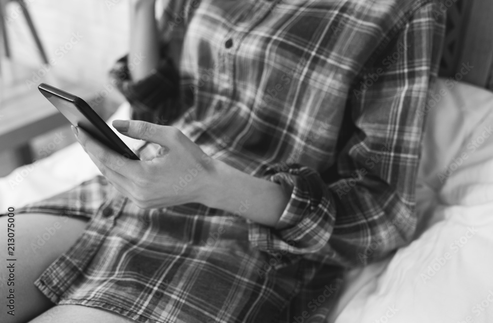 Beautiful girl is sitting on the white bed linen. Young woman in men's checkered shirt is searching in social network on white pillow. Morning with black smartphone in hands.