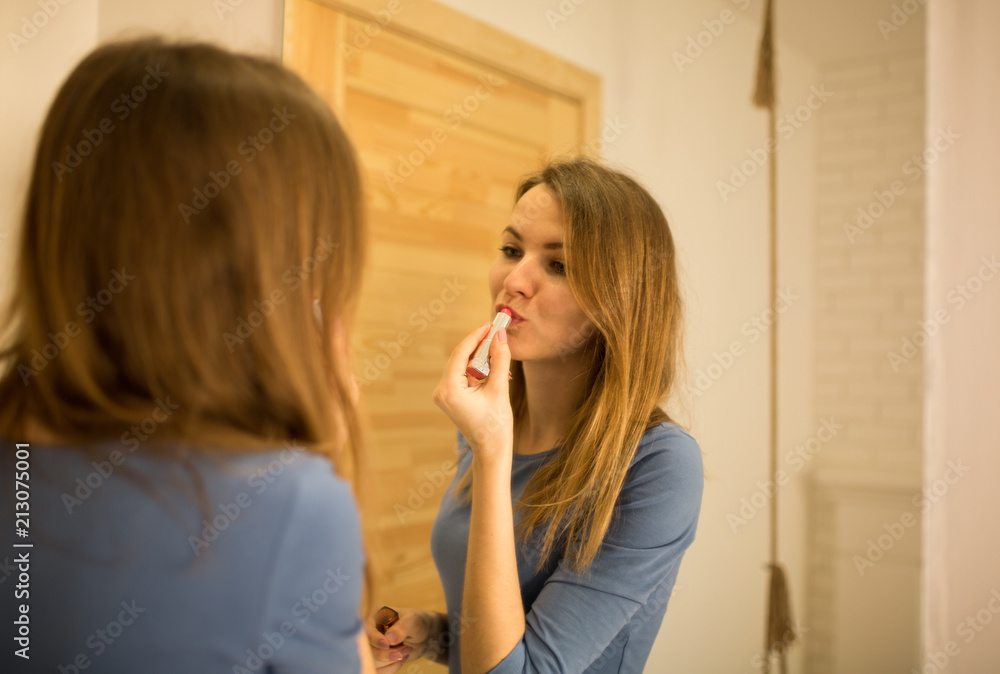 Beautiful girl in blue dress is standing near the big mirror in minimalist loft design room. Young woman is doing make up. She is looking in the mirror and puts on pink lipstick. Beautiful girl in blue dress is standing near the big mirror in minimalist loft design room. Young woman is doing make up. She is looking in the mirror and puts on pink lipstick.