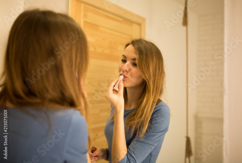 Beautiful girl in blue dress is standing near the big mirror in minimalist loft design room. Young woman is doing make up. She is looking in the mirror and puts on pink lipstick.