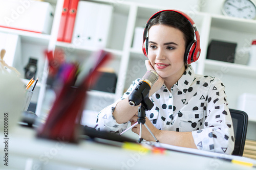 Beautiful young girl sitting in headphones and with microphone at table in office and talking. photo