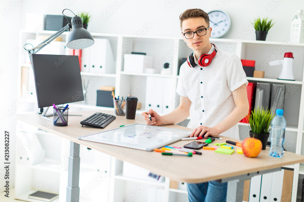 A young man in glasses stands near a computer desk. A young man draws a marker on a magnetic board. On the neck, the guy's headphones hang. A young man in glasses stands near a computer desk. A young man draws a marker on a magnetic board. On the neck, the guy's headphones hang.