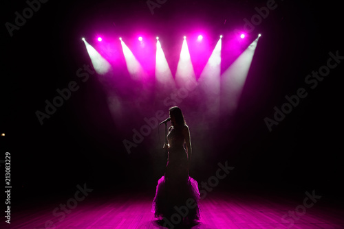 Girl in long gown performing on stage. Girl singing on the stage in front of the lights. Silhouette of singer standing on stage at microphone.