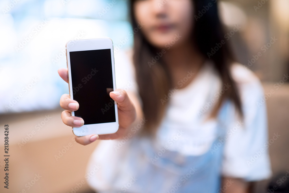 Mockup image of a woman holding and displaying a white mobile phone with a black screen with a blank screen on a table in a modern cafe. Ideas for working with communication tools.