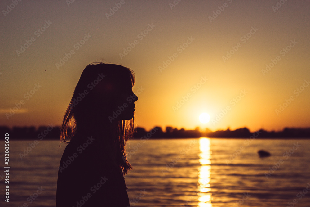 Silhouette of young woman sitting near the water on sand beach with sand in hands