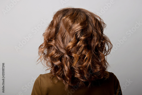 Female hairstyle long curls on the head of a brown-haired woman back view on a gray background.