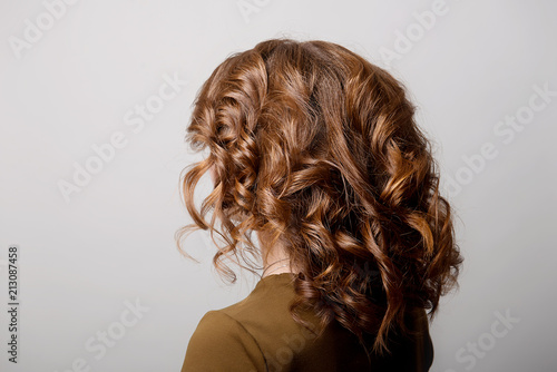 Female hairstyle long curls on the head of the brown-haired woman looking back at the gray background turning the head to the left.