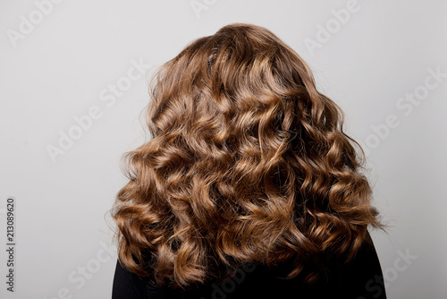 Female hairstyle long curls on the head of a brown-haired woman back view on a gray background.