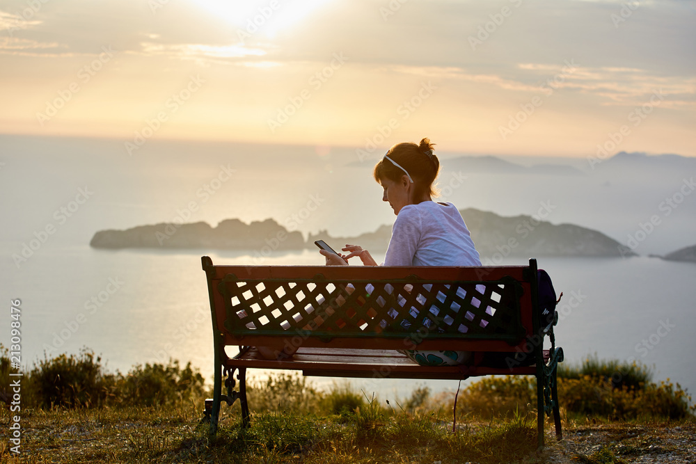 Young woman on a sunset background, the sea and mountains sits on a bench and looks at a smartphone