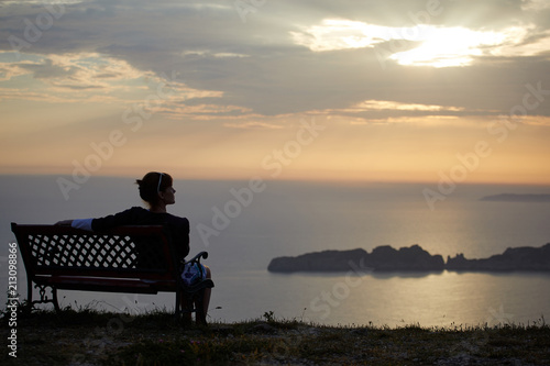Young woman sits on a bench on a sunset background over the sea and mountains