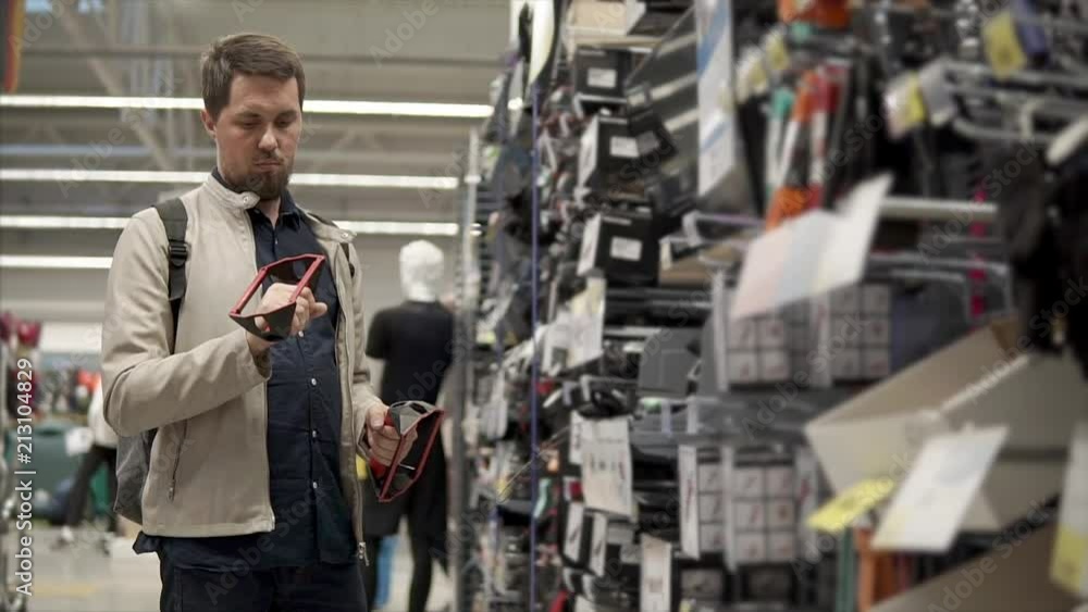 Man is watching on a rack with sport goods in a shop and holding parts of a trainer. He is thinking about buying equipment for exercise for hands Man is watching on a rack with sport goods in a shop and holding parts of a trainer. He is thinking about buying equipment for exercise for hands