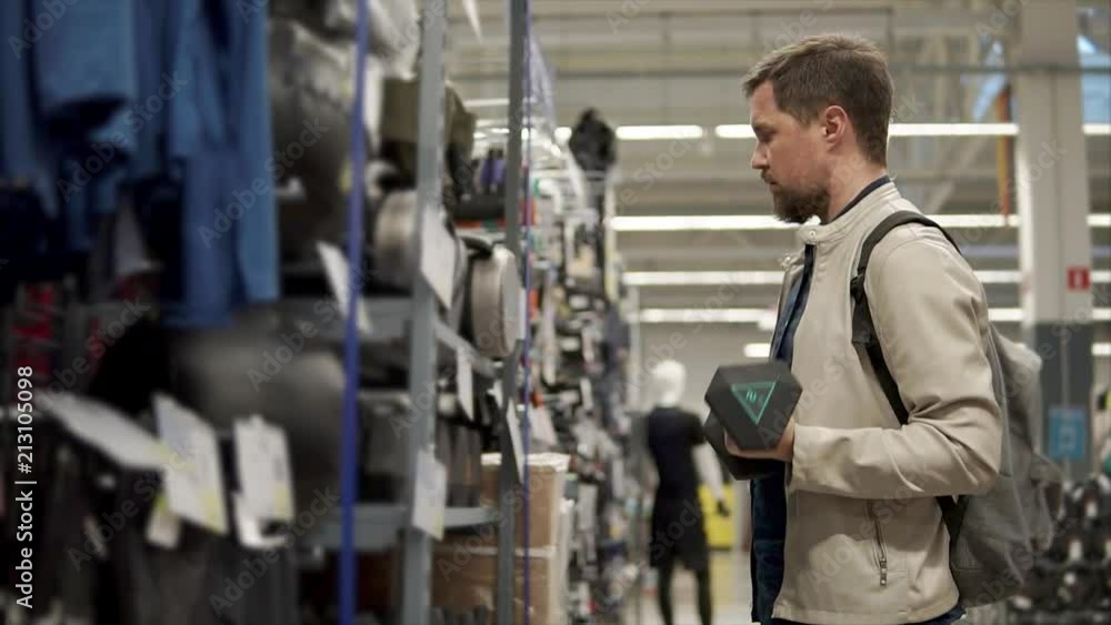 Sporty man is choosing a dumbbell for physical exercises in a shop. He is spinning it in hand and trying on a weight, looking on a shelf